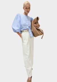 Hellbluse mit weißen Streifen und Puffärmel, kombiniert mit weißen hochtaillierten Hosen und einer geflochtenen braunen Tasche. Schuhe mit Absatz in neutralen Tönen.