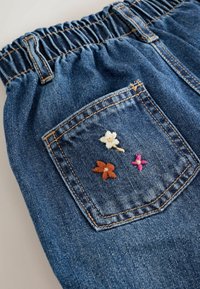 Rückentasche einer blauen Jeanshose mit elastischem Taillenbund, verziert mit kleinen gestickten weißen, braunen und rosa Blumen.