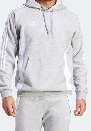 Homme portant un sweat à capuche Adidas gris clair et un pantalon de survêtement assorti avec des bandes blanches et un logo, mains relâchées le long du corps.