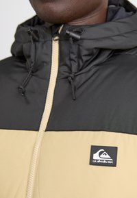 Čierna a béžová prešívaná bunda s kapucňou, ktorá má celý zips a logo Quiksilver na hrudi; nastaviteľné šnúrky na kapucni.