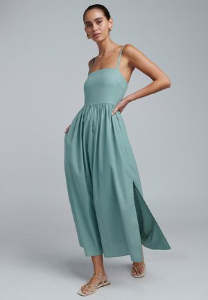 Žena v bezrukávovom skákacom odeve svetlozelenej farby so širokými nohavicami a rozparkami, stojaca s jednou rukou na bokoch a neutrálne výrazom tváre.