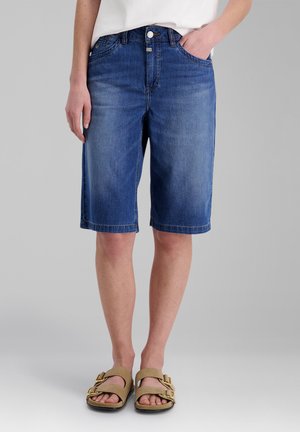 Person wearing mid-length blue denim shorts, white top, and beige sandals with two gold buckles, standing against plain gray background.