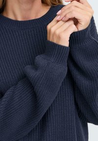 Sötét navy kék kötött pulóver texturált mintával, bordás mandzsettákkal és kerek nyakkivágással, amely laza illeszkedést és puha anyagot mutat.