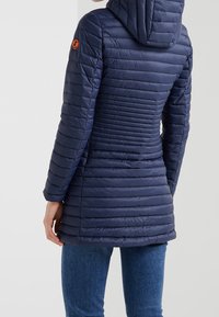 Námořnická puffer bunda s horizontálním prošíváním, kapucí a oranžovou nášivkou na rukávu. Hladká textura, středně dlouhý design.