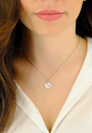 Un collier en argent avec un pendentif en forme de diamant orné d'une image gravée, fixé à une fine chaîne, porté sur une chemise blanche.