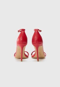 Sandales à talons hauts en cuir rouge avec des brides autour de la cheville, dos ouvert et quincaillerie dorée. Elles présentent une texture lisse et un talon aiguille fin.