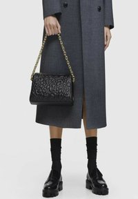 Borsa a tracolla nera trapuntata con catena dorata, caratterizzata da un design testurizzato. Indossata con un soprabito grigio, calze nere e mocassini.