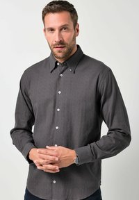 Dunkelgraues Hemd mit Knopfleiste und strukturiertem Muster, mit breitem Kragen und weißen Knöpfen. Das Modell steht mit gefalteten Händen.