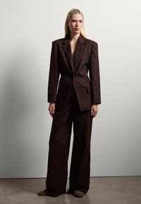 Traje ajustado marrón oscuro con una chaqueta estructurada y pantalones de pierna ancha, hecho de una tela suave, con bolsillos frontales y un escote pronunciado.
