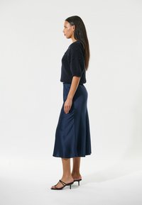 Marineblauer Strickpullover mit strukturierten Puffärmeln, kombiniert mit einem fließenden Satin-Midikleid. Schwarze Riemchen-Heels runden das Outfit ab.