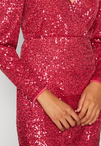 Robe rose pailletée avec manches longues, tissu texturé, décolleté en V profond et bordures roses contrastantes aux poignets. Gros plan sur les mains.