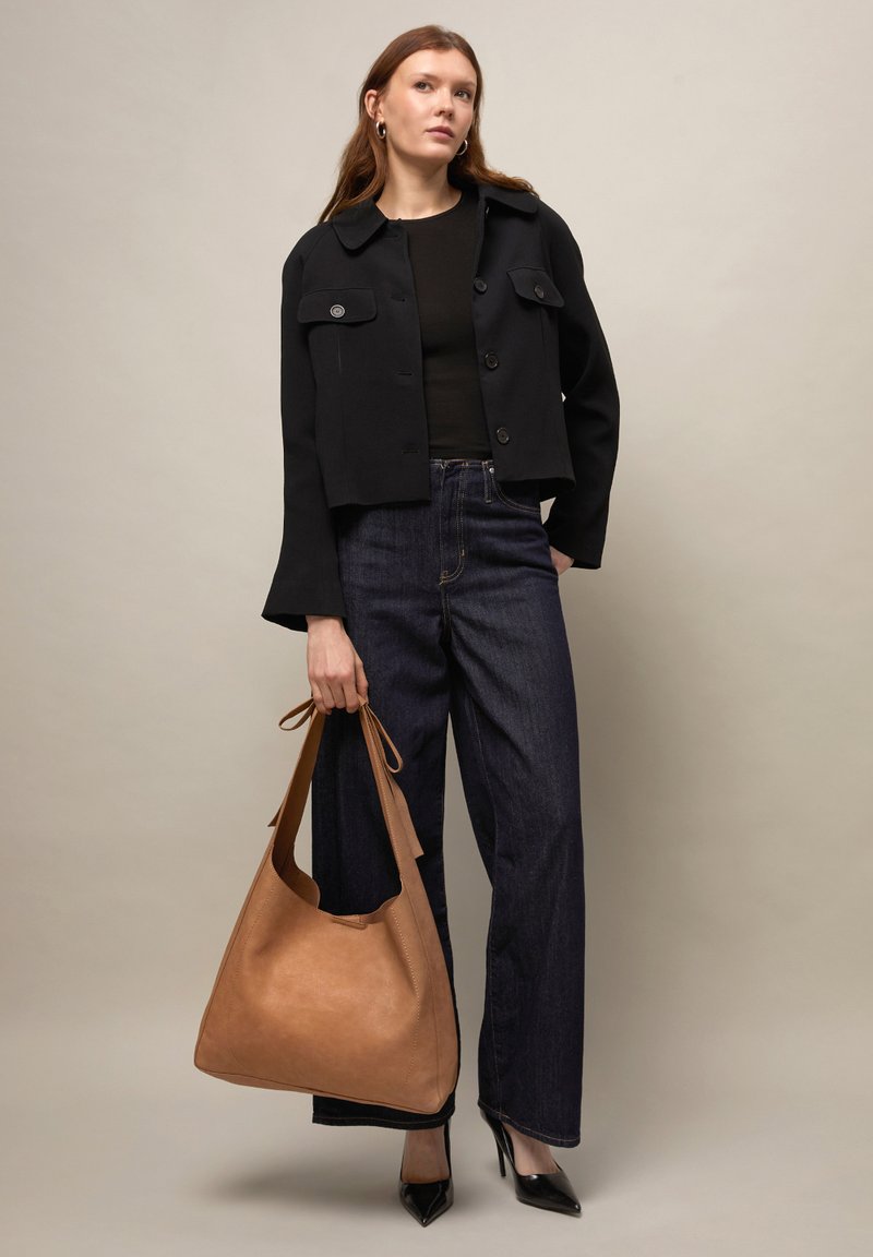 Donna che indossa una giacca corta nera, un top nero, jeans scuri a gamba larga, tacchi neri e tiene una grande borsa in pelle color cuoio.