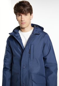 Marinblå vattentät jacka med huva, med en framzipper, knappstängning och två framfickor. Slät textur och lätt design.