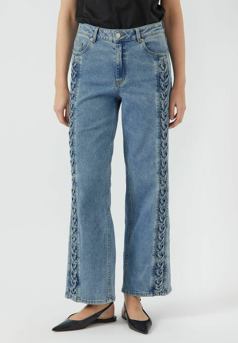 Los pantalones vaqueros de pierna ancha en denim azul presentan detalles trenzados en los costados, cinco bolsillos y un cierre de botón. La tela tiene una textura con un acabado desgastado.
