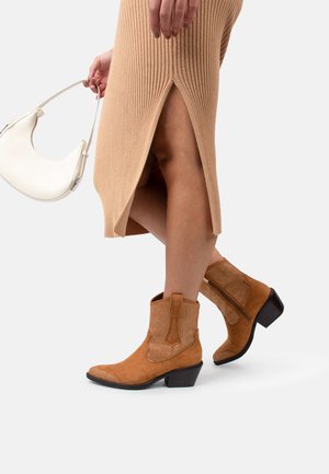 Botines de ante color marrón claro con un tacón bajo, que presentan perforaciones decorativas y costuras en contraste. Se combinan con un vestido beige de canalé.