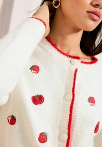 Valge kootud kardigan, millel on punase tomati tikand, kontrastne punane ääris ja ümarad valged nööbid. Pehme tekstuur on nähtav.
