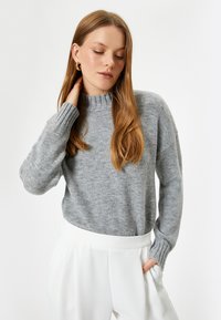 Grauer Strickpullover mit einem gerippten hohen Kragen und Bündchen, der eine entspannte Passform und weiche Textur aufweist. Dazu werden weiße hochgeschnittene Hosen getragen.
