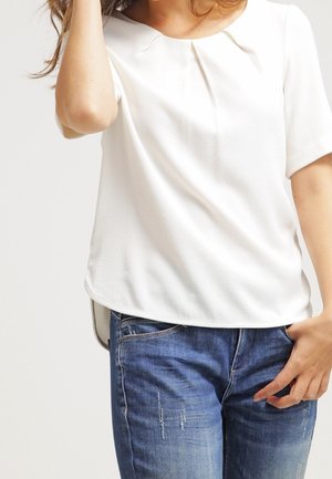 Vrouw draagt een simpele witte blouse met korte mouwen en blauwe distressed jeans, met één hand in haar zak en de andere hand die haar haar aanraakt.