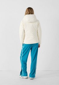 Chaqueta tipo puff en crema con capucha, combinada con pantalones deportivos azules con franjas blancas a los lados y botones a presión en los tobillos.