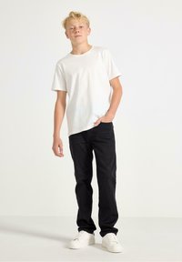 Vit kortärmad T-shirt kombinerad med svarta jeans. Jeansen har rak passform och modellen bär vita sneakers. Enkla bakgrunden.