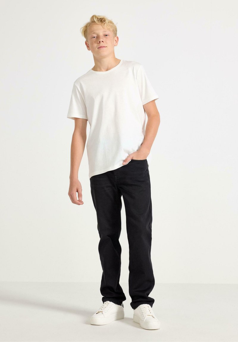 Vit kortärmad T-shirt kombinerad med svarta jeans. Jeansen har rak passform och modellen bär vita sneakers. Enkla bakgrunden.