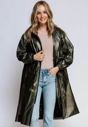 Grüner, glänzender, langer Regenmantel mit elastischen Bündchen und einem Reißverschluss, getragen über einem hellrosa Hemd und blauen Jeans. Glatte, synthetische Textur.