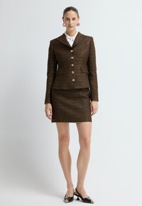 Brązowy garnitur w kratę z dopasowaną marynarką z dużym kołnierzem i złotymi guzikami, w połączeniu z dopasowaną mini spódnicą i czarnymi butami na obcasie typu slingback.