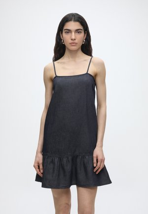 Pikkade tumedate juustega naine, kes kannab varrukateta tumedat teksakleiti, millel on õhukesed rihmad ja volangidega alumine äär, seistes ühevärvilise hele tausta ees.