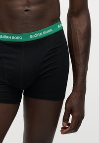 Torse et bras masculins portant un boxer noir Björn Borg avec une ceinture verte affichant le nom de la marque.