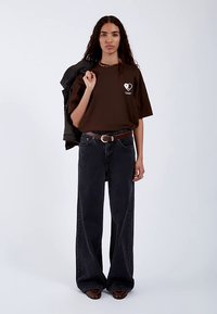 Maglietta oversize marrone con grafica di un cuore bianco, abbinata a jeans a gamba larga neri e giacca di jeans drappeggiata su una spalla.