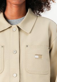 Beige skjorte med knapper og krave, to lommer og et broderet Tommy Jeans logo. Har trykknapper og en glat stofstruktur.