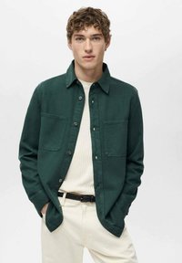Camicia verde scuro con bottoni e due tasche sul petto, indossata sopra un maglione a coste chiaro, abbinata a pantaloni color crema e una cintura nera.
