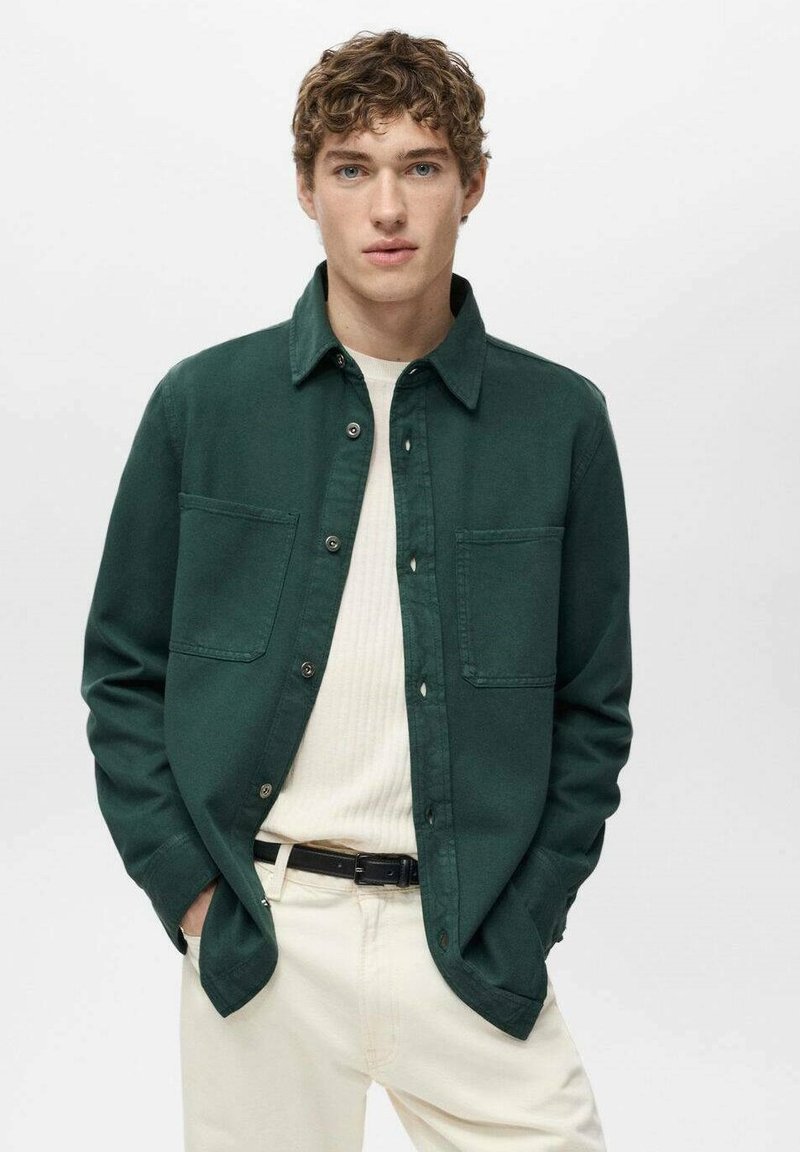 Camicia verde scuro con bottoni e due tasche sul petto, indossata sopra un maglione a coste chiaro, abbinata a pantaloni color crema e una cintura nera.