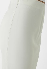 Detailní záběr osoby v těsném světle béžové sukni s vysokým pasem a viditelným bočním švem na jednobarevném bílém pozadí.