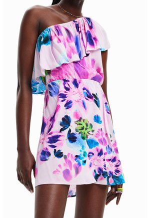 Femme portant une robe colorée asymétrique avec des motifs floraux roses, violets, bleus et verts, ainsi qu'un collier de perles sur fond blanc.