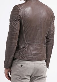 Uomo che indossa una giacca di pelle marrone aderente con pannelli trapuntati sulle spalle e sulle braccia, abbinata a pantaloni beige chiaro, mostrato di spalle.