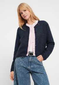 Mørk marineblå, myk cardigan med lange ermer, rund hals og knappeåpning. Brukt over en lys rosa ribbestrikket genser og blå denimjeans.