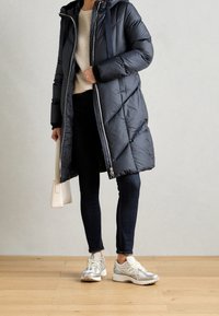 Parka negra acolchada con capucha, que presenta costuras diagonales, un cierre frontal y bolsillos laterales. Llevada sobre un suéter crema y jeans oscuros.