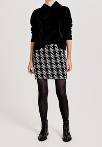 Personne portant un sweat à capuche noir, une jupe pied-de-poule noire et blanche, des collants noirs et des bottes noires à lacets, debout sur un fond gris.