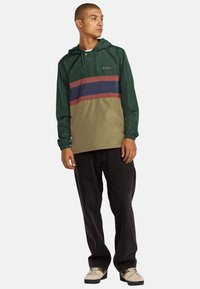 Giacca anorak leggera a strisce verde scuro, beige e bordeaux. Presenta un cappuccio, polsini elastici e una tasca frontale. Texture liscia.