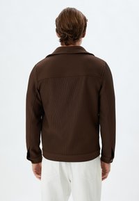 Chaqueta marrón con relieve y cuello, con puños abotonados y corte recto, combinada con pantalones blancos. La tela tiene una apariencia texturizada.