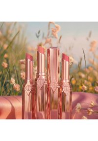 Vier Lippenstifte in Roségold mit verschiedenen Pinktönen, aufrecht stehend in einer blumigen Außenumgebung mit unscharfem Blumenhintergrund.