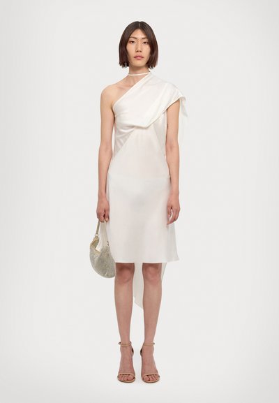 Asymmetrical white dress in lightweight fabric, featuring one shoulder drape, knee-length cut, and a sleek, minimalist design. Accessorized with a small metallic bag.