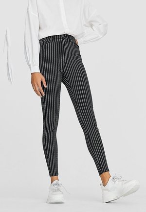 Person wearing black and white vertical striped slim pants with elastic waist, white blouse, and white sneakers standing against a plain background.