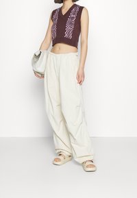Cropped brown sleeveless top with pink woven pattern, paired with loose cream pants and beige slip-on sandals. Holding a light gray handbag.
