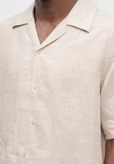 Chemise beige en lin à manches courtes avec col camp et boutons, portée par une personne au teint moyen.