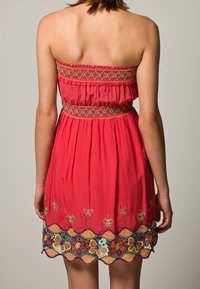 Vestido rojo sin tirantes con una banda bordada en la cintura y un ribete de encaje floral. Presenta patrones coloridos en el dobladillo y una textura de tela fluida.