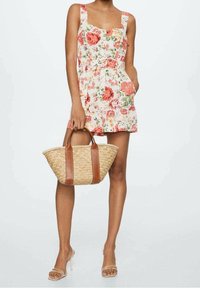 Bloemendress met vierkante halslijn, mouwloos, witte stof met roze en groene bloemen. Houdt een stro bag met bruine leren hengsels vast.