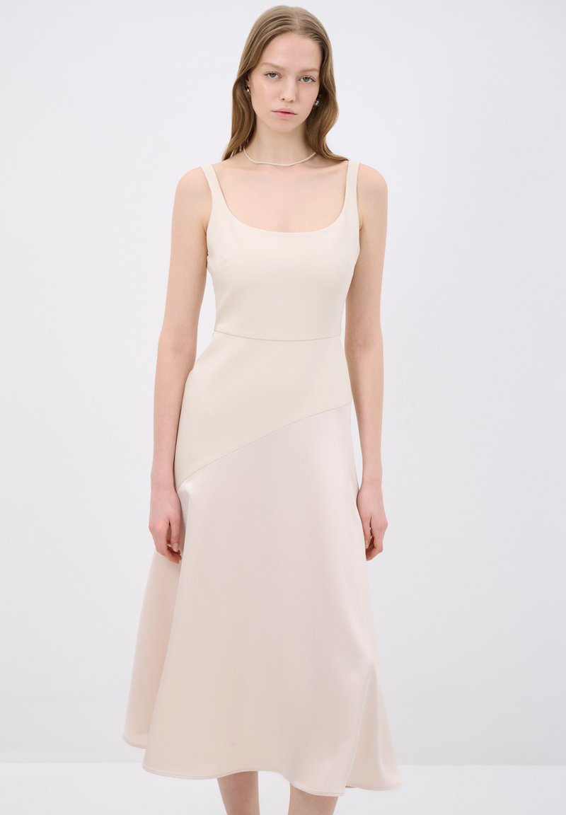 Satin midi-mekko vaaleanbeigellä sävyllä, jossa on neliönmallinen pääntie, istuva yläosa ja liehuva helma epäsymmetrisellä muotoilulla.