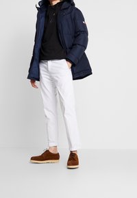Giacca puffer navy con cappuccio, felpa nera sottostante, pantaloni bianchi e scarpe in suede marrone con suole chiare. Senza motivi o accenti visibili.